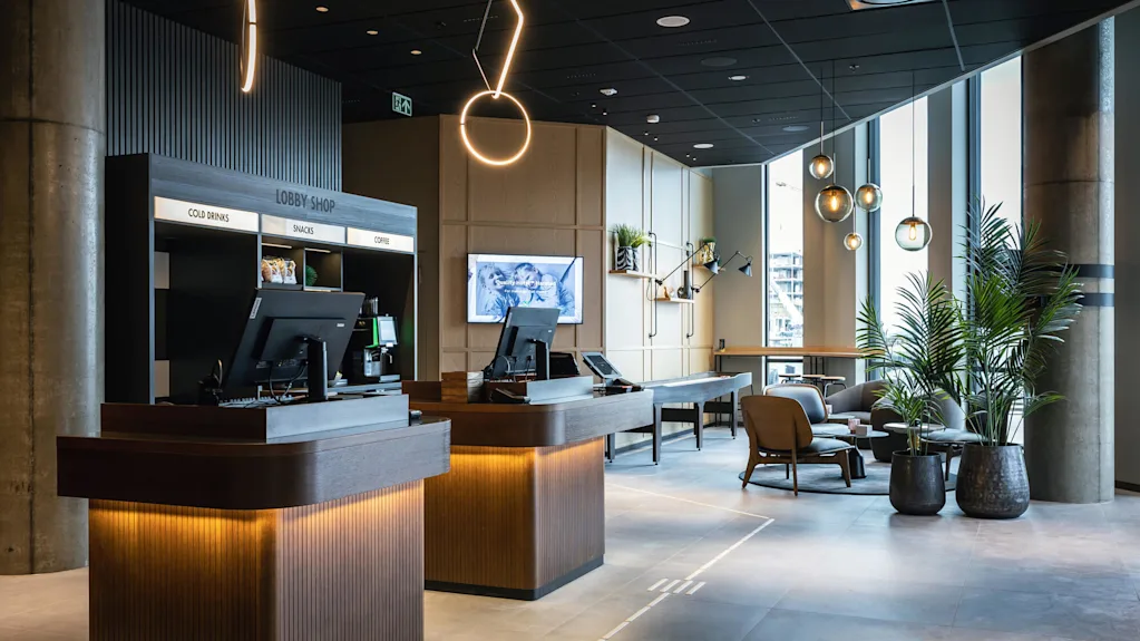 Reception borde står klar i en moderne lobby, oplyst af dekorative lys. Skiltet lyder "LOBBY SHOP: COLD DRINKS, SNACKS, COFFEE". Planter og siddepladser skaber en indbydende atmosfære.