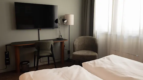 Et koselig hotellrom med en seng, et funksjonelt skrivebordsområde med stol, en veggmontert TV og en komfortabel lenestol ved et vindu.