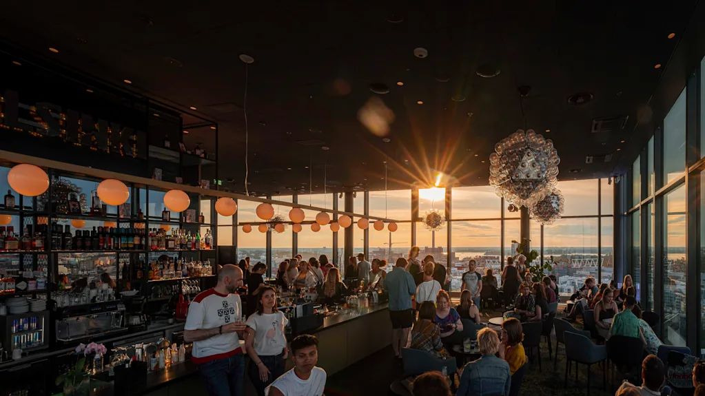 Barbesökare njuter av solnedgång genom stora fönster, med glödande sfäriska lampor ovanför. Helsingfors skyline syns och "HELSINKI" är upplyst på en hylla.