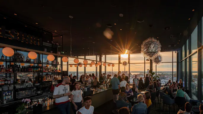 Barpatroner nyter solnedgangsutsikt gjennom store vinduer, med glødende sfæriske lys over hodet. Helsingfors skyline er synlig, og "HELSINKI" er opplyst på en hylle.