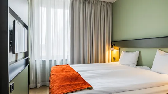 Et koselig hotellrom med en pent oppredd seng med hvite laken og et oransje pledd. Myk belysning fra nattbordslamper gir varme. En flatskjerm-TV er montert på veggen. Store vinduer er dekket med gardiner.