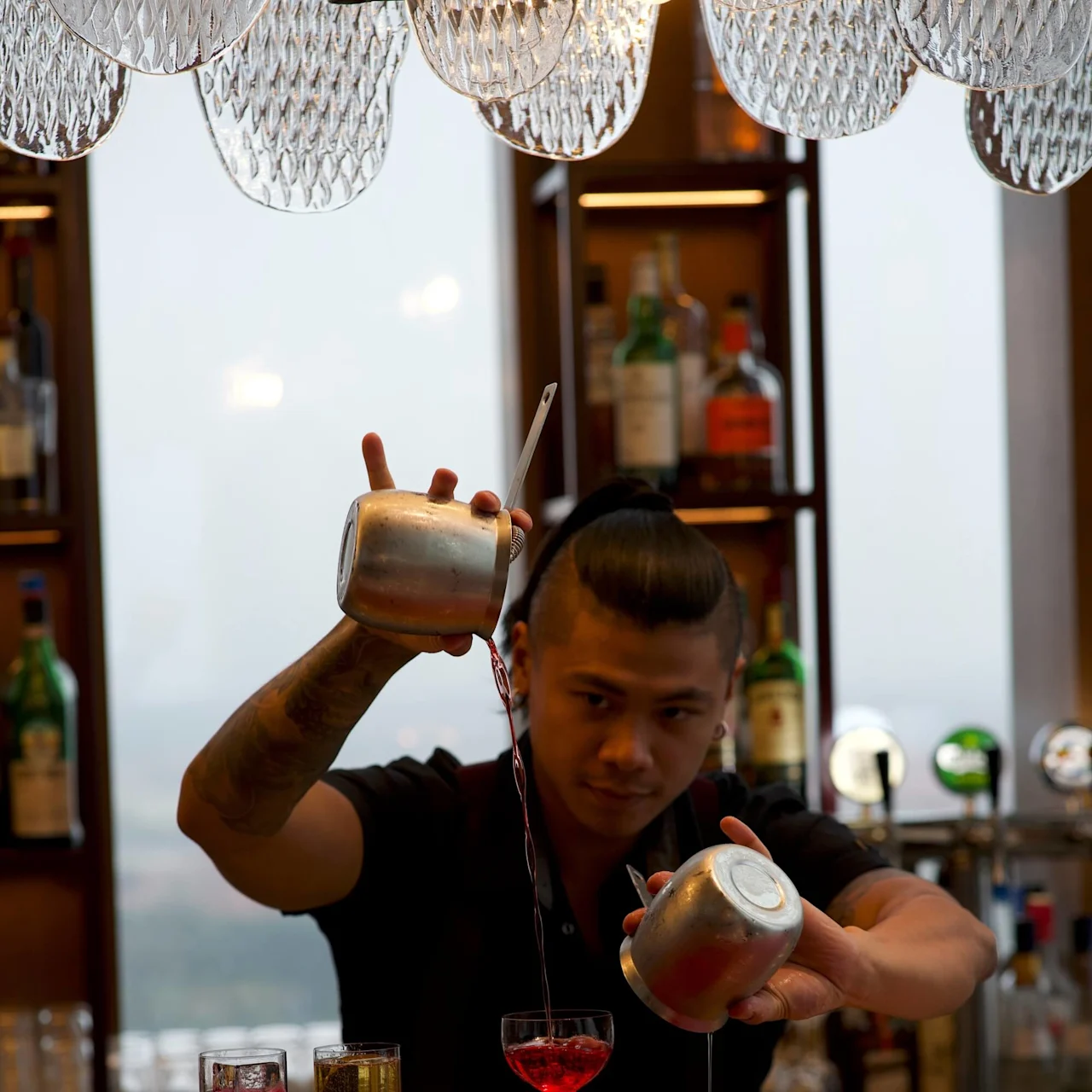 Bartender skjenker dyktig drikke i glass fra to metallrister og er fokusert. Baromgivelser med forskjellige flasker og glass på hyller i bakgrunnen.