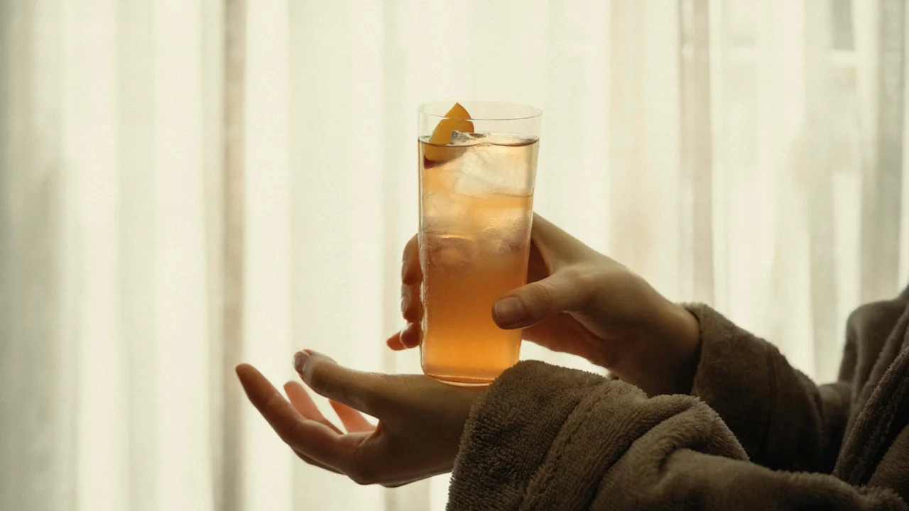 Person in a bathrobe holding a refreshing drink with ice and a citrus garnish, relaxing by a window.