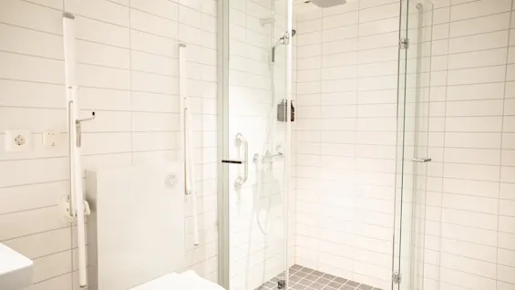 Toalett placerad bredvid en dusch med glasdörrar och moderna beslag, omgiven av vita kaklade väggar och ett grått klinkergolv, vilket skapar en ren, minimalistisk badrumsmiljö.