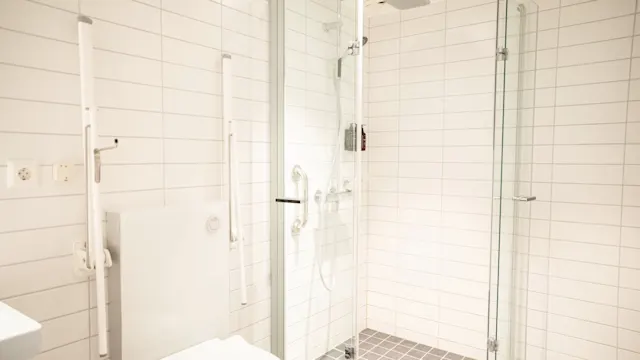 Toalett plassert ved siden av en dusj med glassdører og moderne beslag, omgitt av hvite flislagte vegger og et grått flislagt gulv, som skaper en ren, minimalistisk baderomssetting.