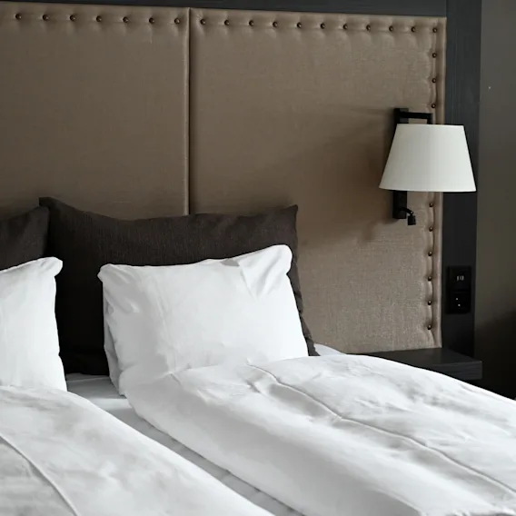 A neatly made bed with white pillows and a duvet, set against a beige upholstered headboard. A wall-mounted lamp is on the right beside the bed.
