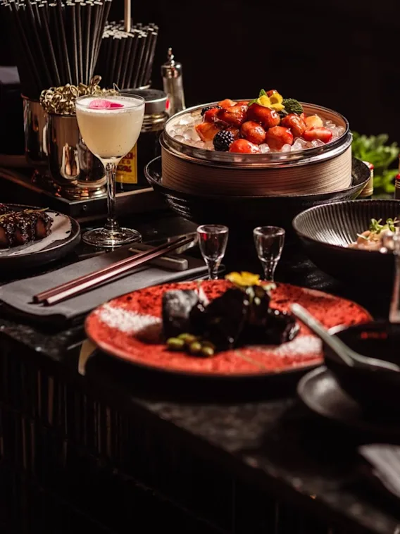 En mängd olika rätter och drycker är arrangerade på ett svagt upplyst restaurangbord, med frukt på is, en cocktail och små tallrikar med garneringar, omgivna av bestick och glas.