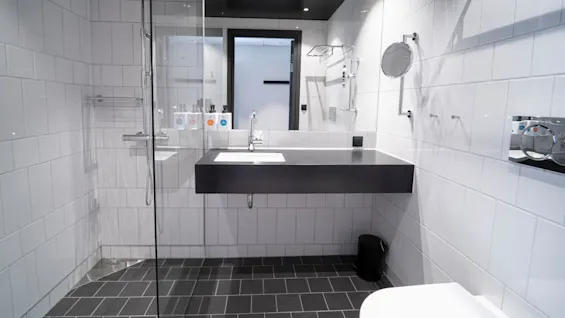 A modern bathroom features a sleek black countertop with a sink, large mirror, and glass shower. White tiled walls and dark grey floor enhance the minimalist design.