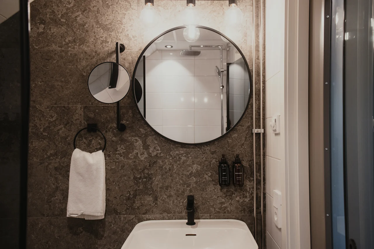 Ett modernt badrum med en rund spegel ovanför ett vitt handfat, med mörka texturerade plattor och en fräsch handduk.