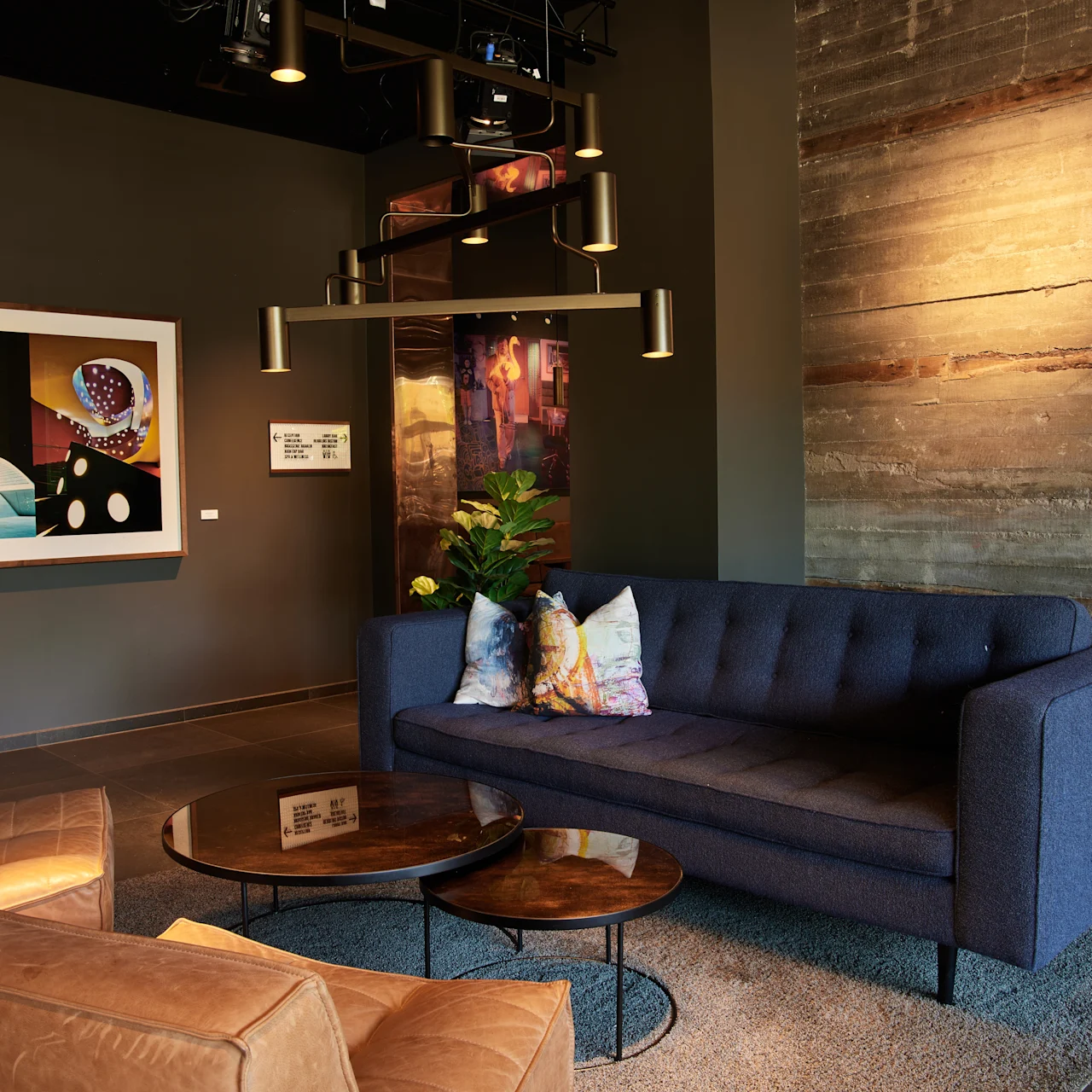 Blå soffa står i en modern lounge med bruna läderfåtöljer och runda träbord. Konst och växter pryder rummet, belyst av en modern takarmatur.
