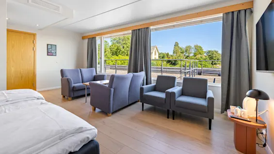 A modern hotel room with a bed, seating area, and open balcony door, showcasing a sunny outdoor view.