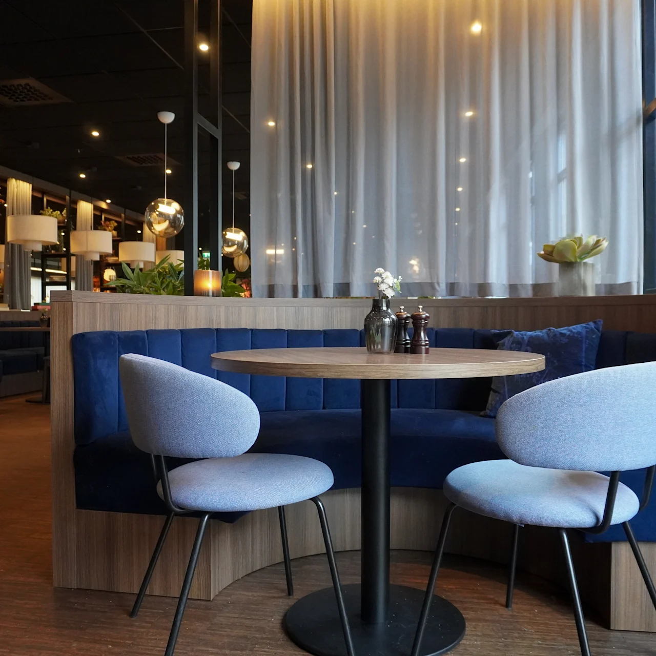 Ett runt träbord med två grå stolar står framför en blå stoppad bänk i en mysig, välbelyst restaurang med skira gardiner och modern inredning.