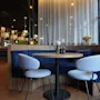 Ett runt träbord med två grå stolar står framför en blå stoppad bänk i en mysig, välbelyst restaurang med skira gardiner och modern inredning.