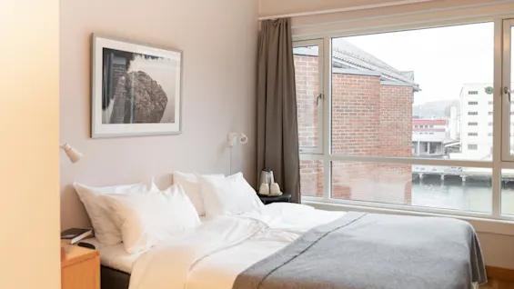 Et lyst og innbydende hotellrom med en pent oppredd seng, et innrammet kunstverk og et stort vindu med utsikt over vannet.