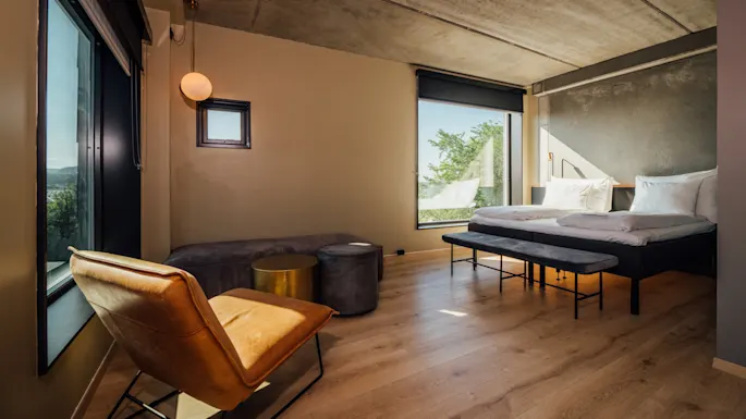 Bedroom with a modern bed, two pillows, and a blanket. A tan chair, round stool, and small table sit by large windows. Natural light fills the room with greenery visible outside.