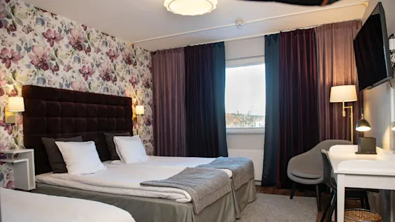 A hotel room features two white-covered beds against a floral wallpaper, with a dark headboard. Nearby, a desk with a lamp and chair sits beneath a wall-mounted TV.