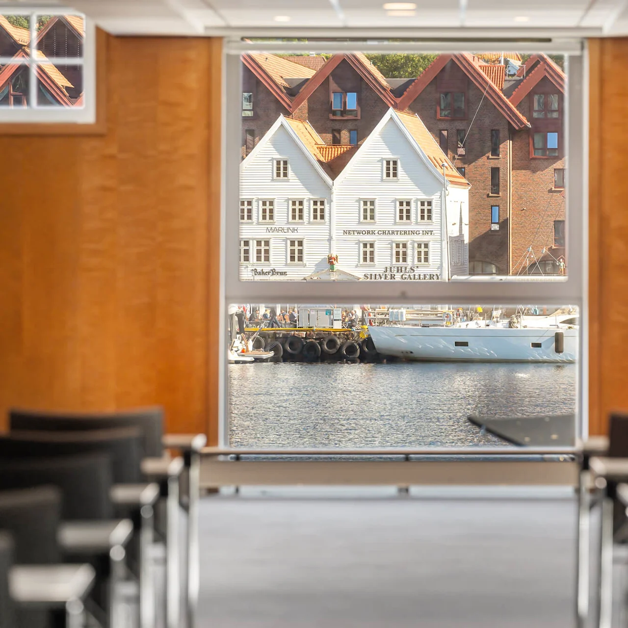 Konferencerummets stole vender mod et stort vindue med udsigt over en havn, både og farverige bygninger. Tekst inkluderer "Marunik," "Network Chartering Inc." og "Hege’s Silver Gallery."