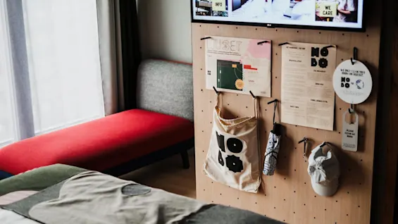 Pegboard with hanging bag, umbrella, cap, and notes; TV above; red bench and bed nearby. Notes read: "HUSET," "WE ONLY CLEAN YOUR ROOM ON REQUEST," "INFO," "PRACTICAL STUFF," "THE BREAKFAST," "HOMO FAB." 