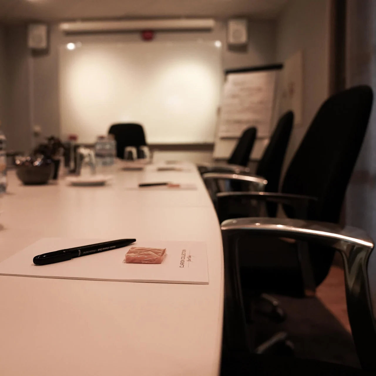 A conference room setup with a long table, chairs, note papers, pens, bottled water, and a projected screen at the front.