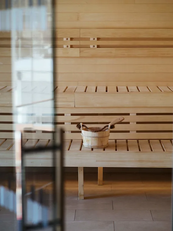A wooden sauna bucket with a ladle rests on a bench inside a wooden sauna, viewed through a glass door.