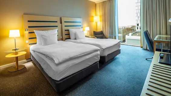 Två prydligt bäddade enkelsängar står sida vid sida, omgivna av lampor, i ett modernt hotellrum med stort fönster, skrivbord och stol.