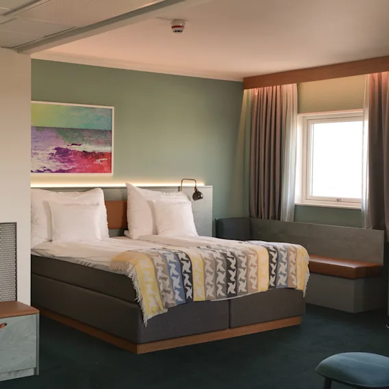 A neatly made double bed with patterned blankets stands in a stylish room featuring a green wall, framed art, and a small window seat bathed in soft natural light.