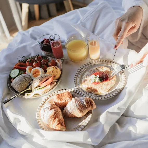 A person in a bathrobe enjoying a delicious breakfast in bed with a variety of food and drinks, bathed in natural light.