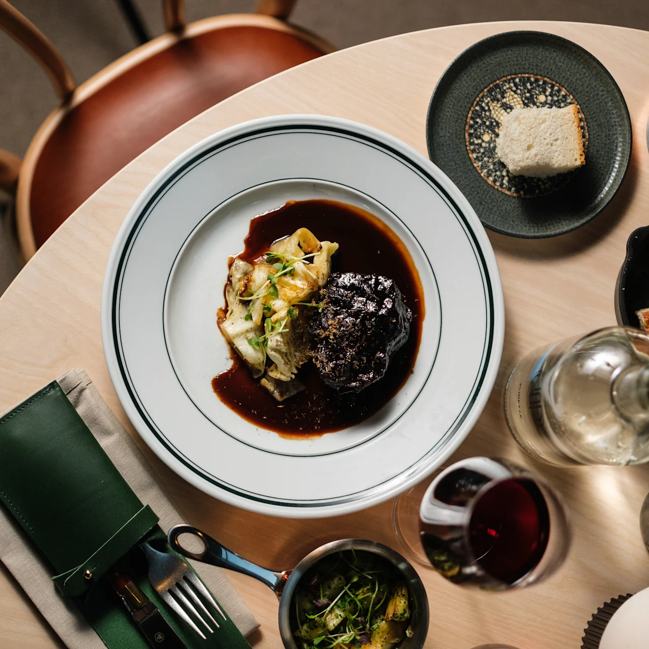 Ett bord med en huvudrätt bestående av kött och grönsaker, omgiven av bröd, smör, grönsaker, ett glas rödvin och en vattenflaska i en matsituation.