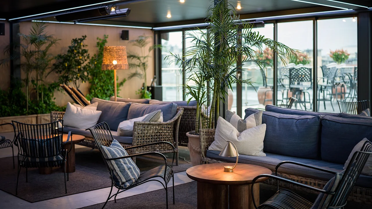Koselig sittegruppe med kurvstoler og sofaer med puter, omgitt av potteplanter, under et moderne tak med varm belysning, ved siden av store vinduer med utsikt over et utendørsområde.