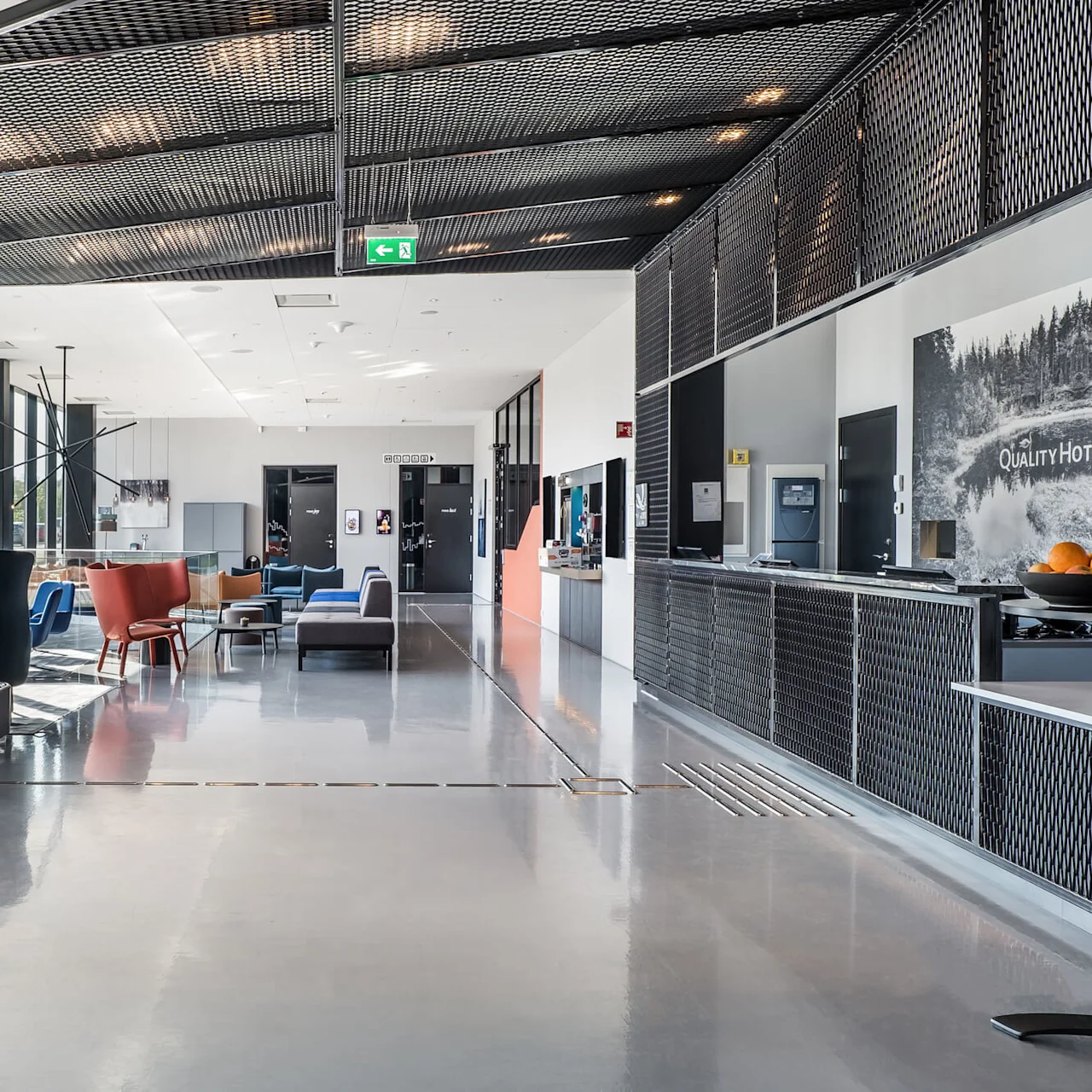A modern hotel lobby features a seating area with colorful chairs and a reception desk. "Quality Hotel" is visible on the wall. Glass doors lead outside, creating a spacious environment.