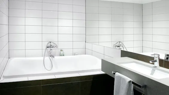 White bathtub with a stainless steel faucet next to a modern sink and large mirror in a tiled bathroom.