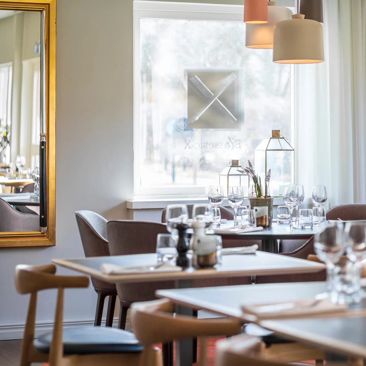 Eleganta middagsbord är dukade med vinglas och servetter under mjuka pendelbelysningar, omgivna av moderna stolar i en ljus, fönsterbelyst restaurang. Text på fönstret: "Brasserie X".