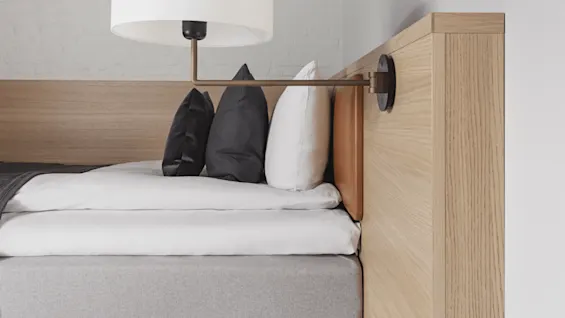Bed with grey and white bedding, black and white pillows, beneath a wall-mounted lamp. Located in a room with a wooden headboard and light-colored walls.