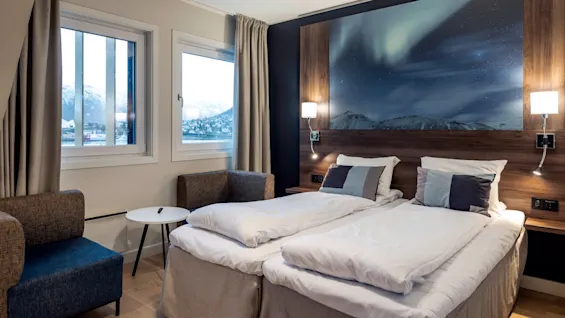 A neatly made double bed with white linens, framed by bedside lamps and an aurora-themed artwork. Chairs and a small table by the window overlook snowy mountains.
