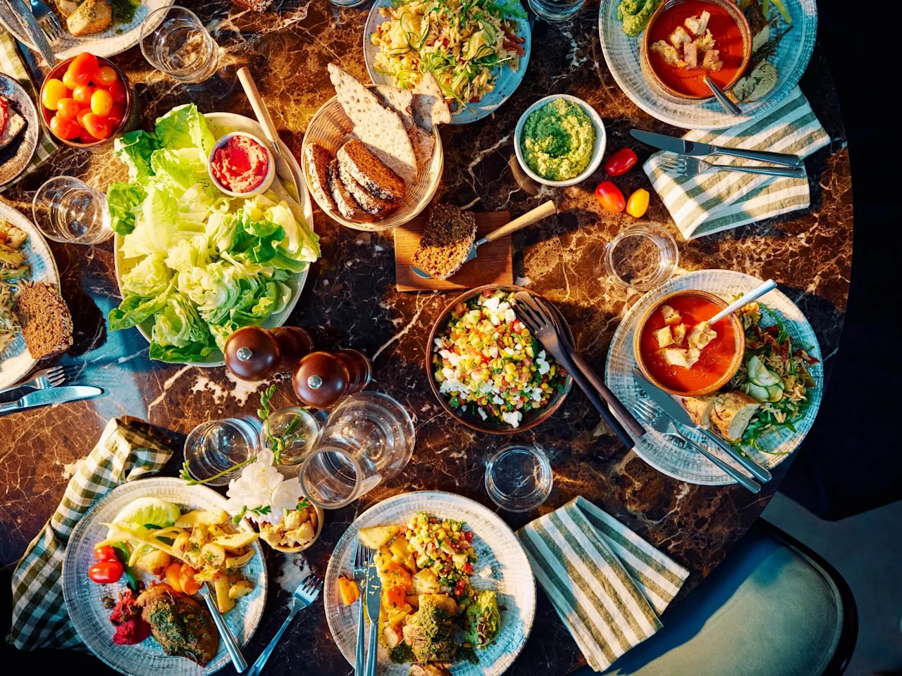 Skåle og tallerkener fyldt med forskellige salater, supper og brød er arrangeret på et mørkt marmorbord, omgivet af bestik, servietter og glas, skaber en farverig middagsopsætning.