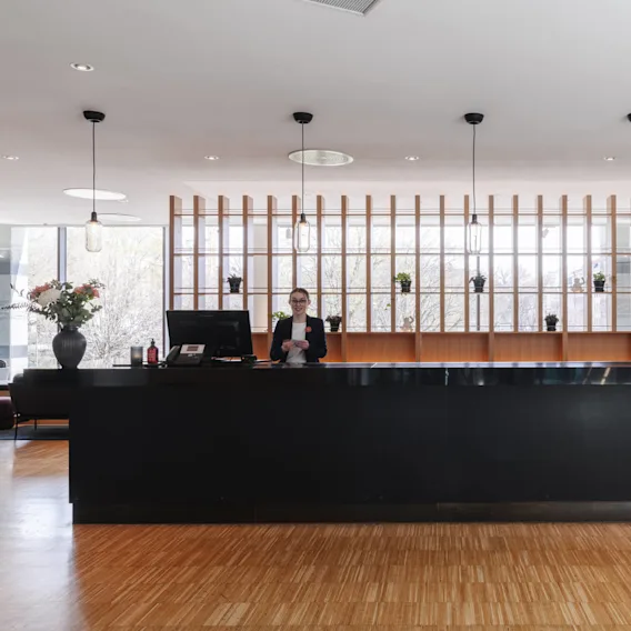 Receptionen bemannas av en leende person i en ljus, modern lobby. Trähyllor med växter pryder bakgrunden, och hängande lampor hänger från taket.