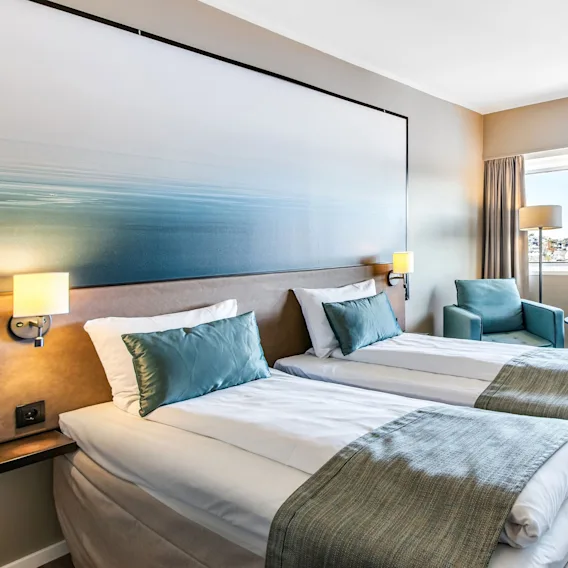 Lyst hotellrom med to enkeltsenger, blå puter og et stort veggmaleri over sengen.