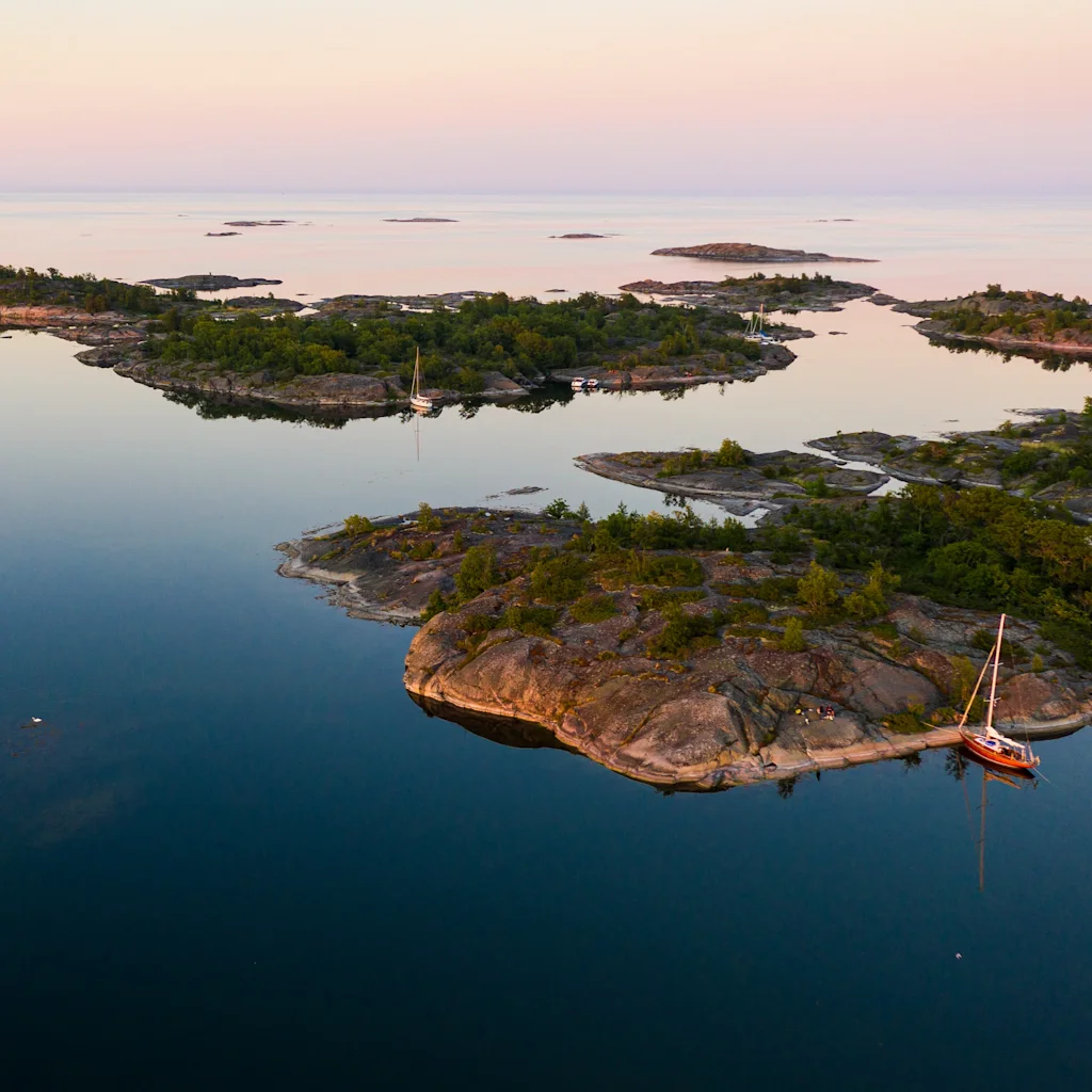 Aerial view of the Stockholm archipelago at dusk, with numerous small, tree-covered islands and sailboats anchored in calm waters.