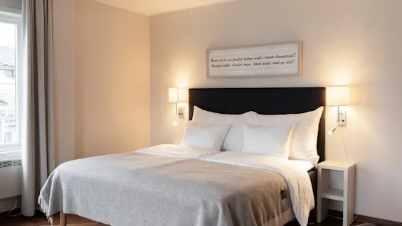 A comfortable hotel room with a bed, soft lighting, and a framed quote: Fuad vil du om frugten sponge midt i træets blomstertid?