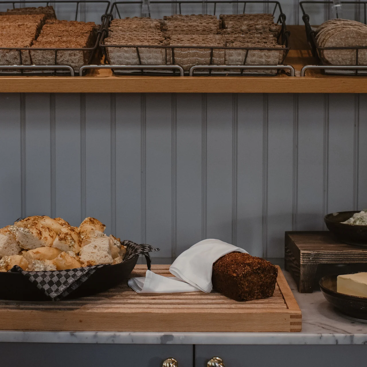 Bröd och bullar ligger på en skärbräda med smör och en limpa bredvid. Ovanför finns hyllor med olika typer av knäckebröd i en köksmiljö.