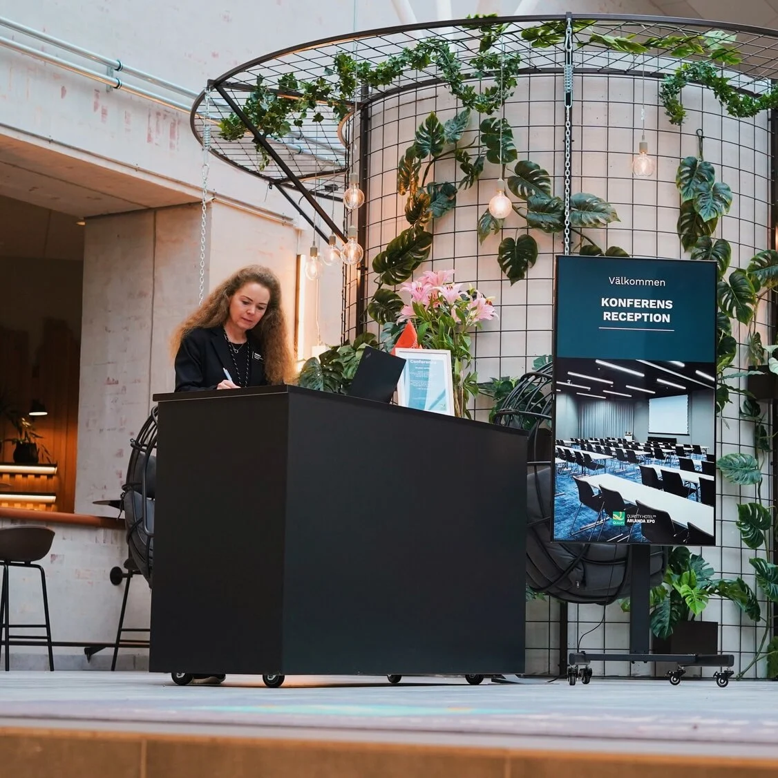 A woman writes at a black counter in a reception area adorned with hanging plants and light bulbs. Nearby, a sign reads "Välkommen KONFERENS RECEPTION" with a conference room image.