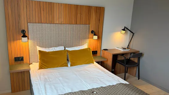 En seng med sennepsfargede puter står mot et trepanel; ved siden av er et skrivebord med lampe og stol på et lyst tregulv.