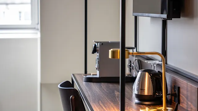 A wooden desk features a silver electric kettle, a coffee maker, and a stylish lamp. A chair is positioned in front, with a window view in the background.