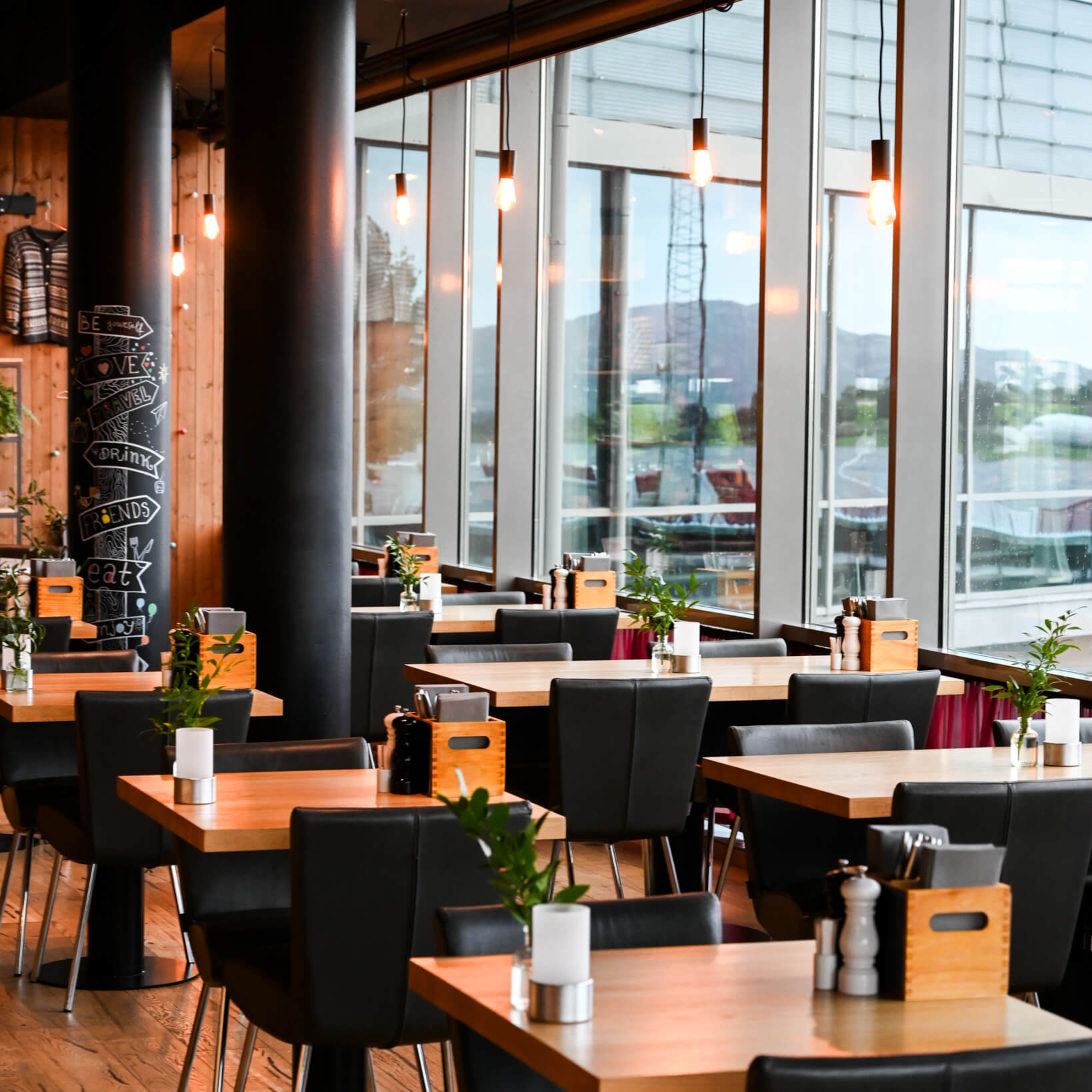 En moderne restaurantindretning med træborde, sorte stole, pendellamper og store vinduer, der giver naturligt lys.