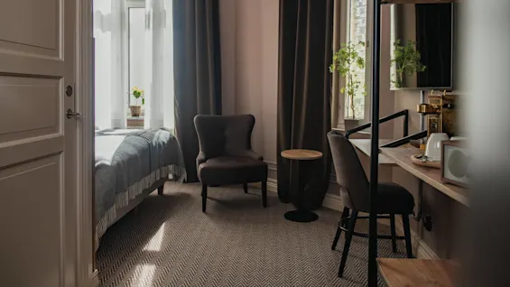 A small, sunlit room features a bed with a gray throw blanket, a cozy armchair, a small side table, and a workspace with a chair and potted plants on windowsills.