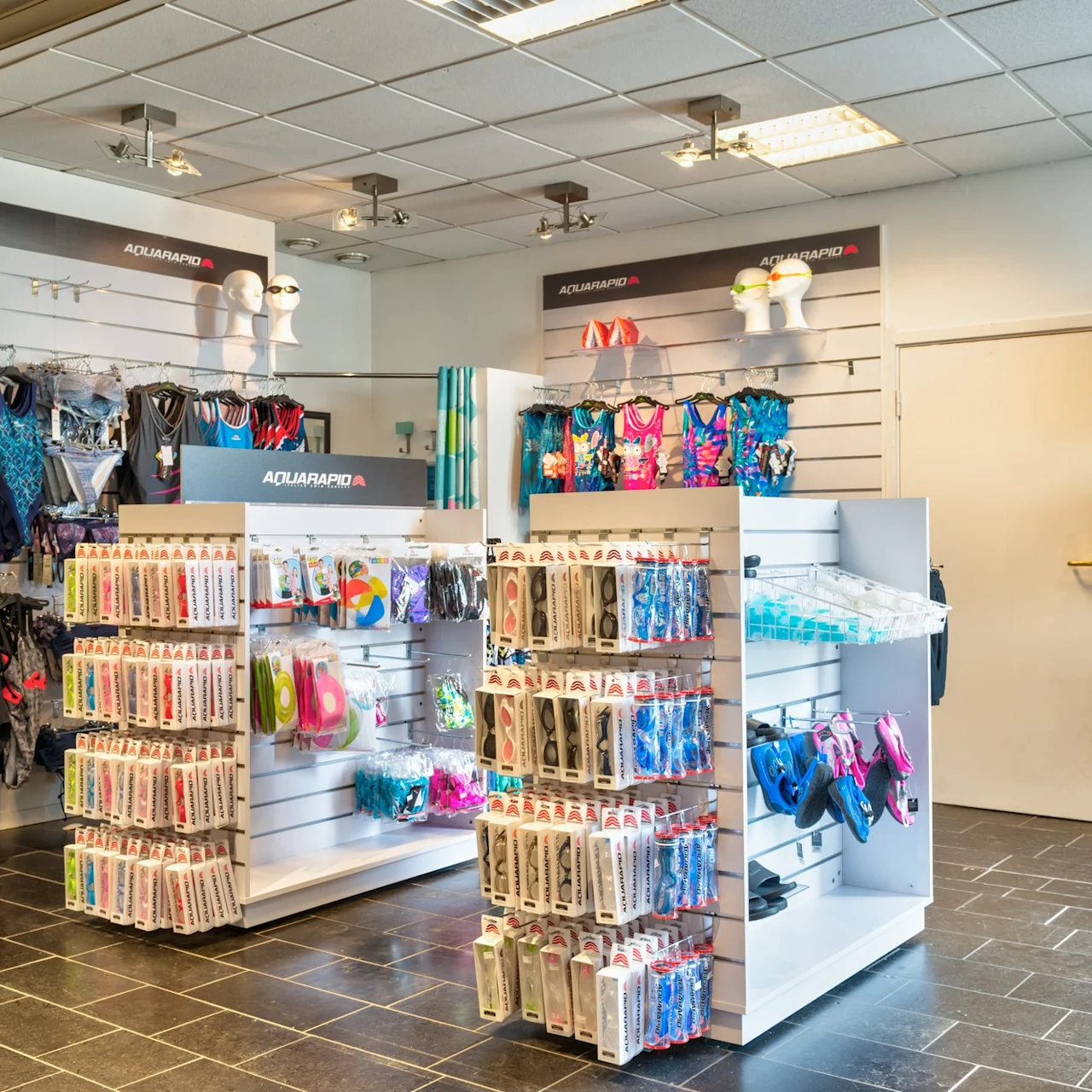 A retail display showcases swimwear and accessories, including goggles and swimsuits. Mannequin heads model goggles. Signage says "AQUARAPID." The bright, organized store has tiled flooring.