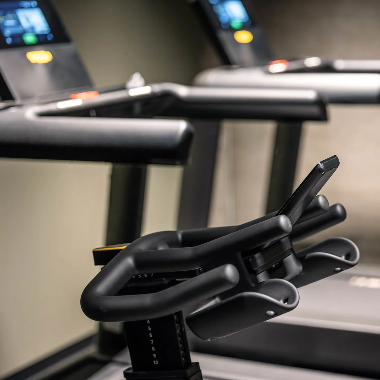 Løbebånd med digitale skærme står stationære i et fitnesscenter. Forgrunden viser et nærbillede af håndtag og en smartphone-holder. Baggrundens skærme er lidt uskarpe.