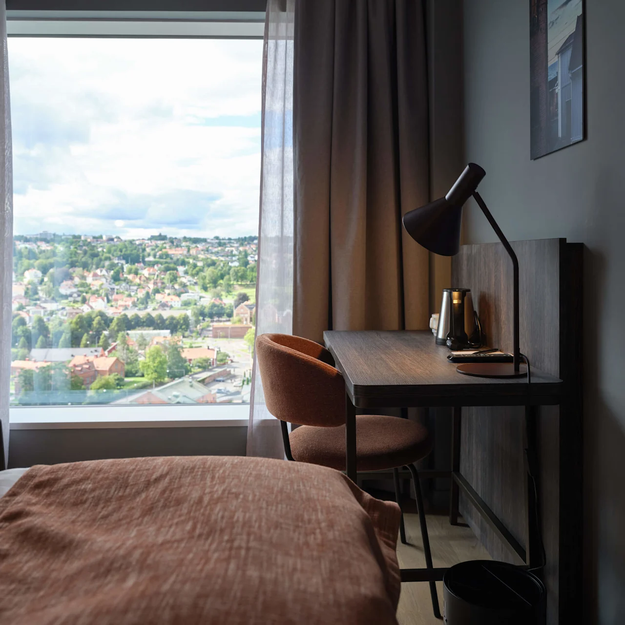 Skrivebord med en lampe og en elkedel står ved siden af et vindue med udsigt over en by. En stol står ved skrivebordet, og en seng er tæt på.