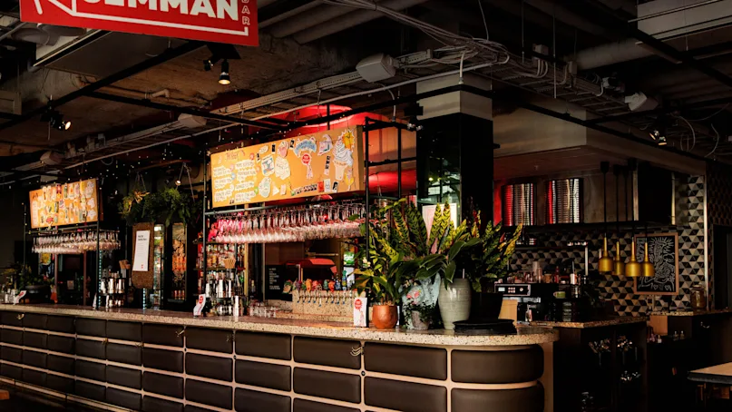 A bar with a curved counter displaying hanging wine glasses and potted plants. Bright orange signage with colorful illustrations hangs above. The setting is a modern, industrial-style space.