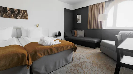 Two neatly made twin beds with brown blankets, a white robe on one, placed in a modern hotel room with a sofa, desk, and large window. Neutral colors enhance tranquility.